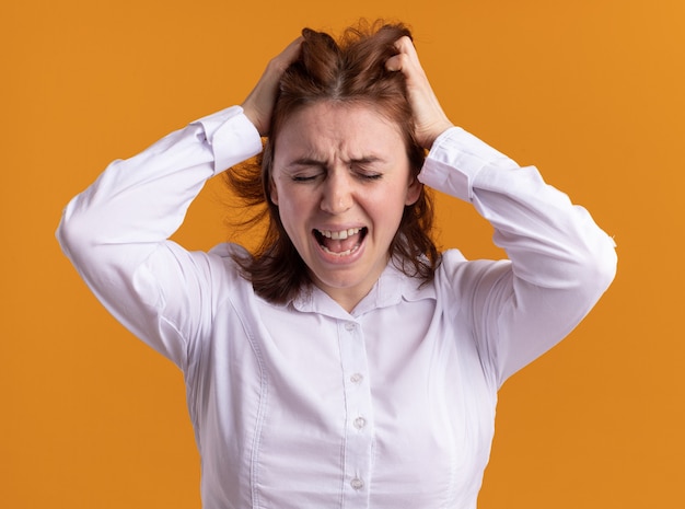 Young woman in white shirt shouting going wild pulling her hair standing over orange wall