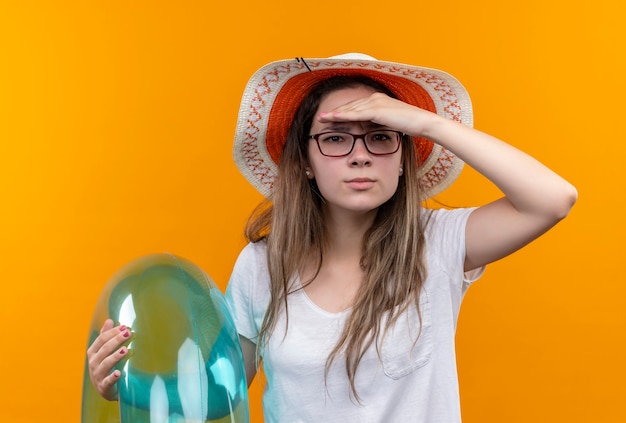 Free photo young woman in white t-shirt wearing summer hat  holding inflatable ring looking far away with hand over head to look someone or something standing over orange wall