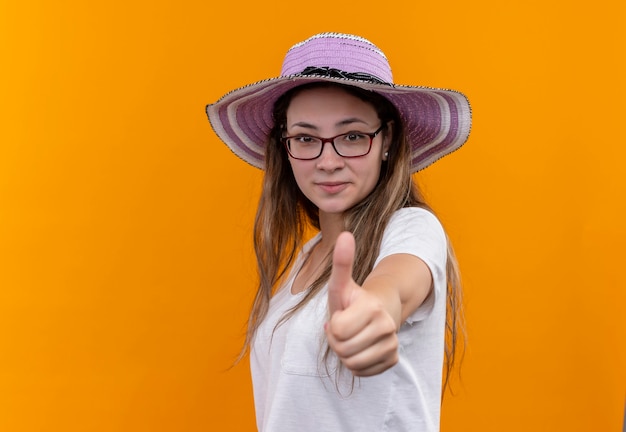 Young woman in white t-shirt wearing summer hat smiling cheerfully showing thumbs up standing over orange wall