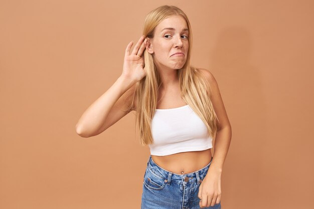 Young woman in white top and blue jeans keeping hand at her ear, listening attentively