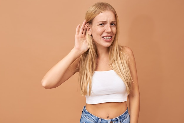 Young woman in white top and blue jeans keeping hand at her ear, listening attentively