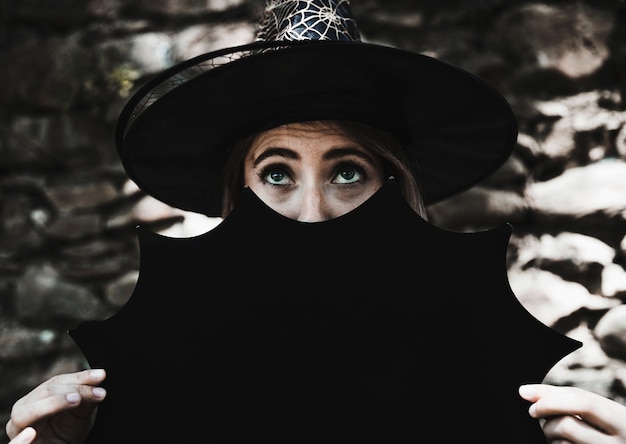 Free photo young woman in witch hat holding halloween decoration near face