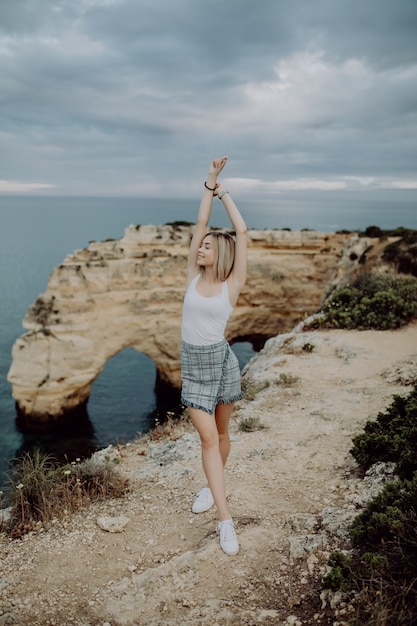 Free photo young woman with arms wide open standing on rock in front of ocean beach on portugal