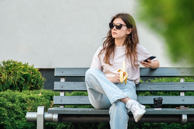 A young woman with a banana sits on a bench and uses a smartphone