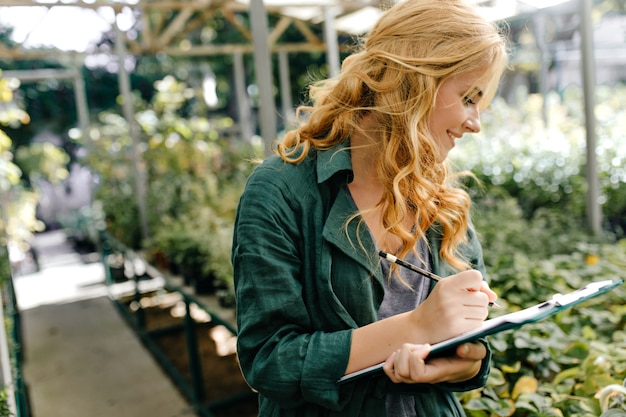 Free photo young woman with beautiful blond hair and gentle smile, dressed in green robe with belt is working in greenhouse