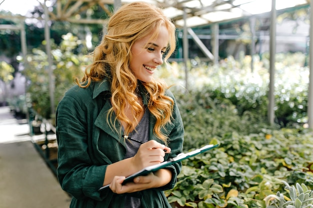 Free photo young woman with beautiful blond hair and gentle smile, dressed in green robe with belt is working in greenhouse