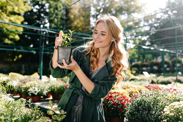 Young woman with beautiful blond hair and gentle smile, dressed in green robe with belt is working in greenhouse