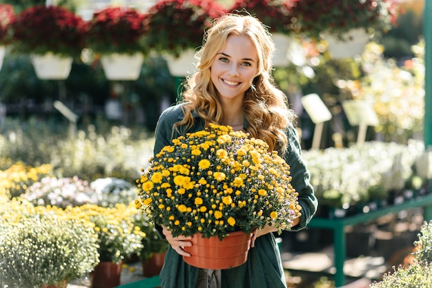 Young woman with beautiful blond hair and gentle smile, dressed in green robe with belt is working in greenhouse