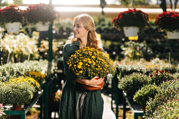 Young woman with beautiful blond hair and gentle smile, dressed in green robe with belt is working in greenhouse