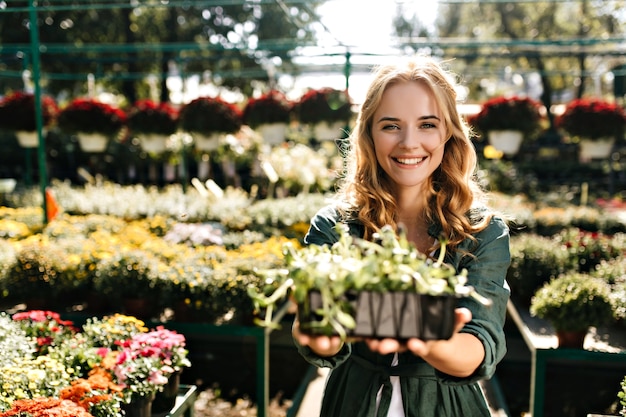 Young woman with beautiful blond hair and gentle smile, dressed in green robe with belt is working in greenhouse