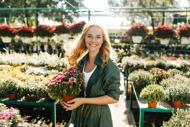 Young woman with beautiful blond hair and gentle smile, dressed in green robe with belt is working in greenhouse