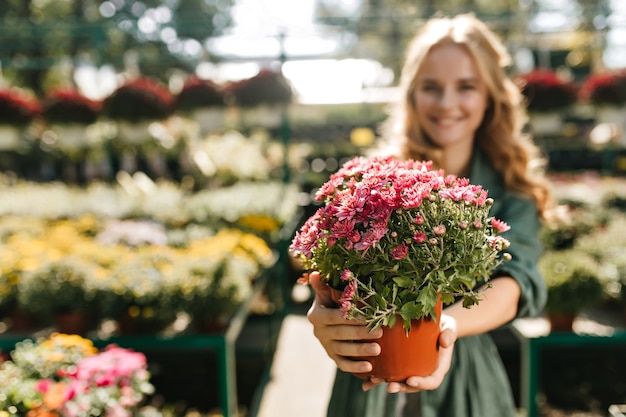 Young woman with beautiful blond hair and gentle smile, dressed in green robe with belt is working in greenhouse