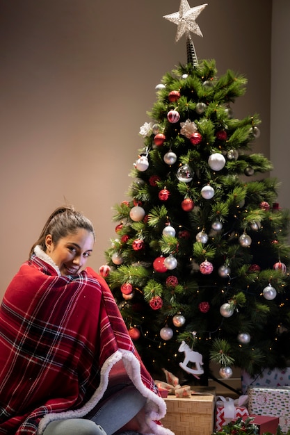 Young woman with blanket sitting next to christmas tree