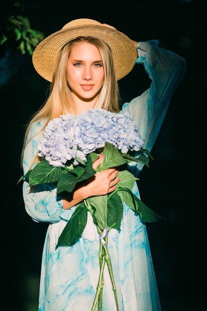 Young woman with blue hedragea buquet in blue dress standing in green garden on sunset