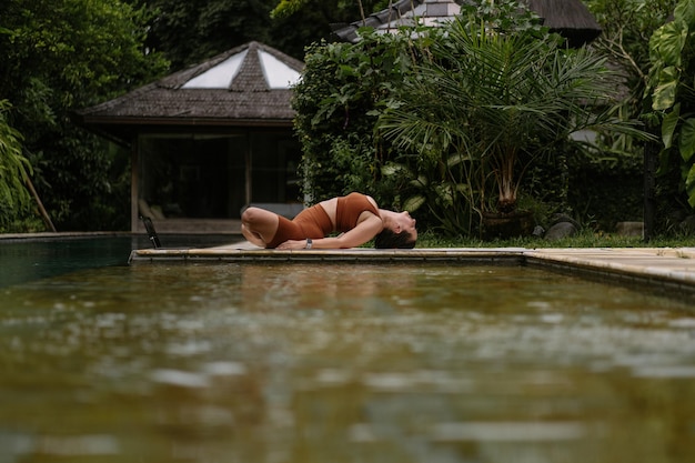 Free photo young woman with body positive appearance practicing yoga alone on deck by the pool in tropical island of bali, indonesia. sport, fitness, healthy lifestyle concept.