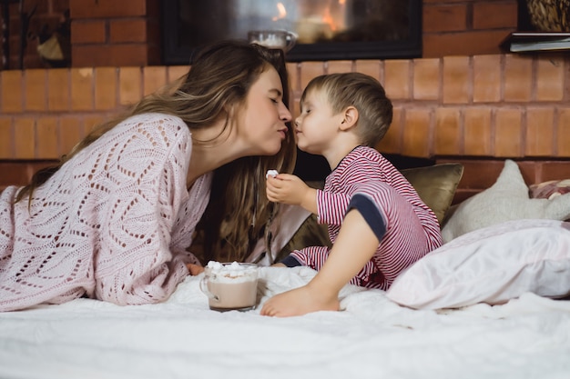 young woman with a child by the fireplace. mom and son drink cocoa with marshmallows 