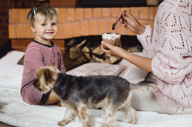 Free Photo young woman with a child by the fireplace. mom and son drink cocoa with marshmello near the fireplace.