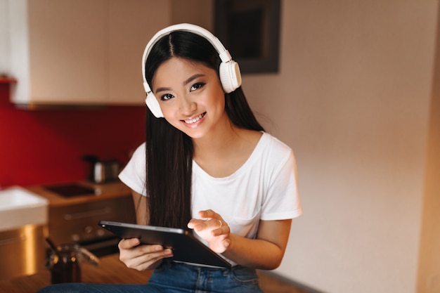 Young woman with dark hair with smile looks into front, holds tablet and listens to music with headphones