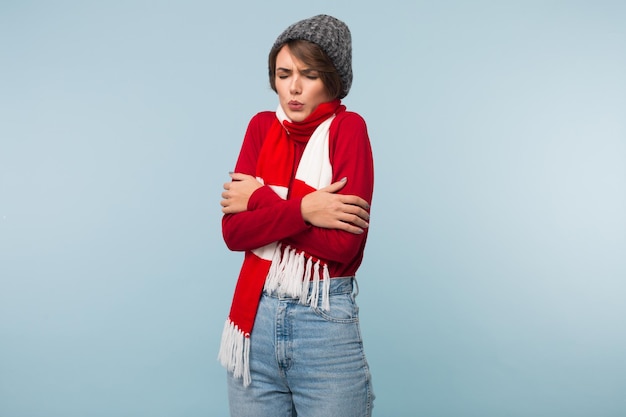 Free Photo young woman with dark short hair in red sweaterscarf and knitted hat shaking from cold over blue background isolated