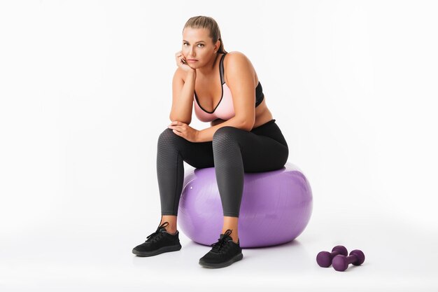 Young woman with excess weight in sporty top and leggings sitting on fitness ball with dumbbells near thoughtfully looking in camera over white background isolated