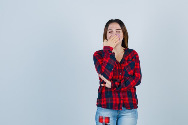 Young woman with hand on mouth in shirt, jeans and looking merry , front view.