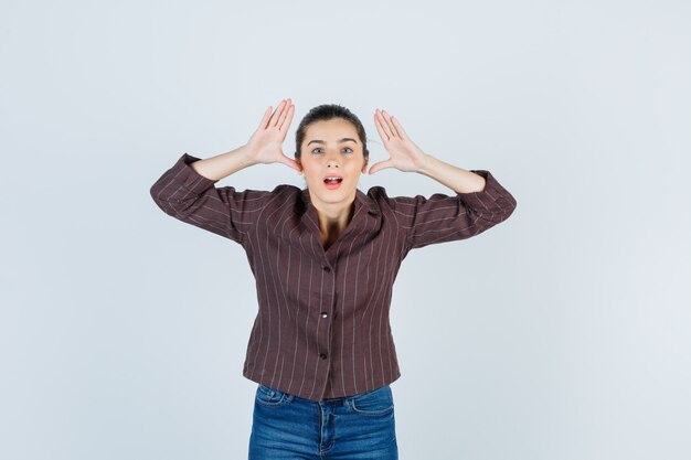 Young woman with hands near head, keeping mouth open in striped shirt, jeans and looking surprised , front view.