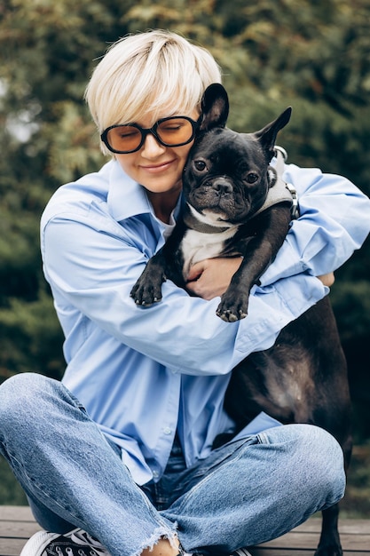 Free photo young woman with her pet french bulldog sitting on a bench in a park