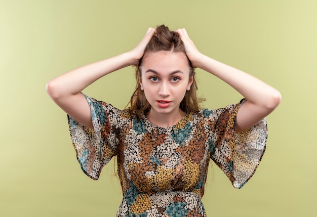 Young woman with long hair wearing colorful dress stressed and frustrated touching head  standing over green wall