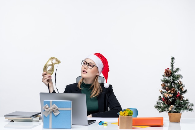 Free photo young woman with santa claus hat and eyeglasses and holding mask sitting at a table with a xmas tree and a gift