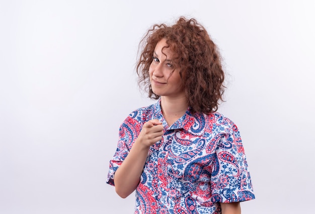 Free photo young woman with short curly hair  in colorful shirt looking confident smiling pointing with finger at front