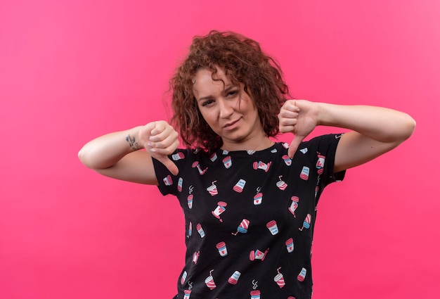 Free photo young woman with short curly hair looking displeased showing thumbs down with both hands standing over pink wall