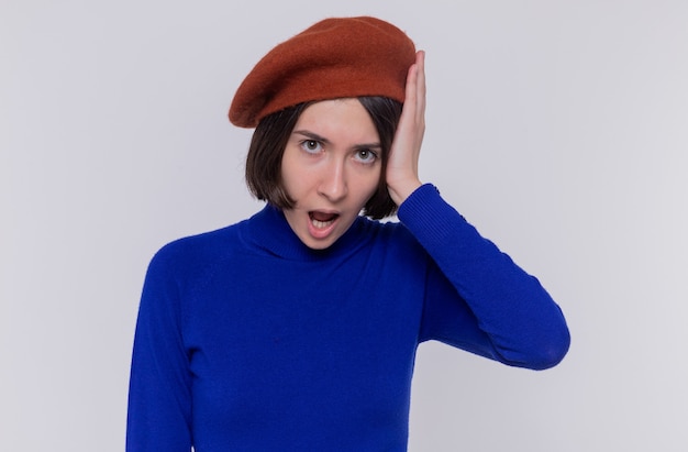 Young woman with short hair in blue turtleneck wearing beret looking at front amazed and surprised standing over white wall