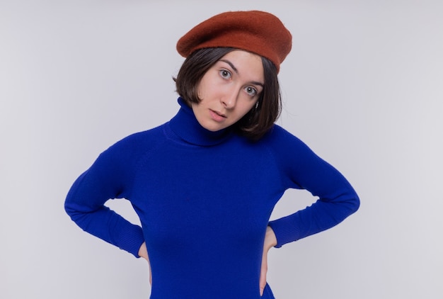 Free photo young woman with short hair in blue turtleneck wearing beret looking at front with skeptic smile on face standing over white wall