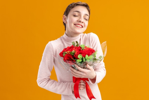 young woman with short hair holding bouquet of red roses happy and positive with eyes closed smiling valentines day concept standing over orange wall