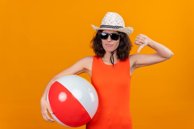 A young woman with short hair in an orange shirt wearing sun hat and sunglasses holding inflatable ball showing thumbs down 