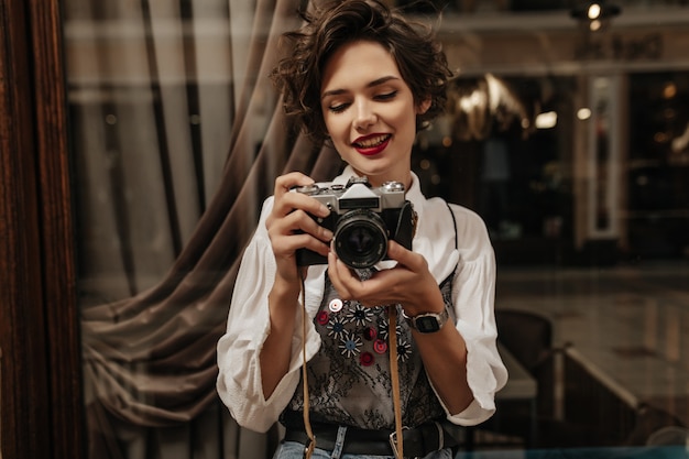 Young woman with short hair and red lipstick making photo inside. Positive woman in stylish white shirt holding camera in cafe.