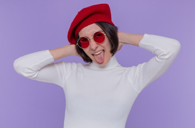 Free Photo young woman with short hair in white turtleneck wearing beret and red sunglasses looking at front happy and joyful sticking out tongue standing over blue wall