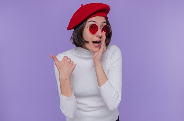 Young woman with short hair in white turtleneck wearing beret and red sunglasses looking surprised telling a secret holding hand near mouth pointing back with thumb standing over blue wall