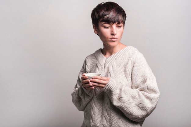 Young woman in woolen sweater holding coffee cup against gray background