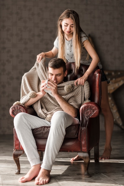 Young woman wrapping her boyfriend with shawl sitting on armchair drinking coffee