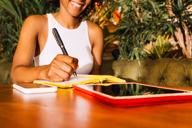 Young woman writing on diary with pen over the wooden table with cellphone and digital tablet