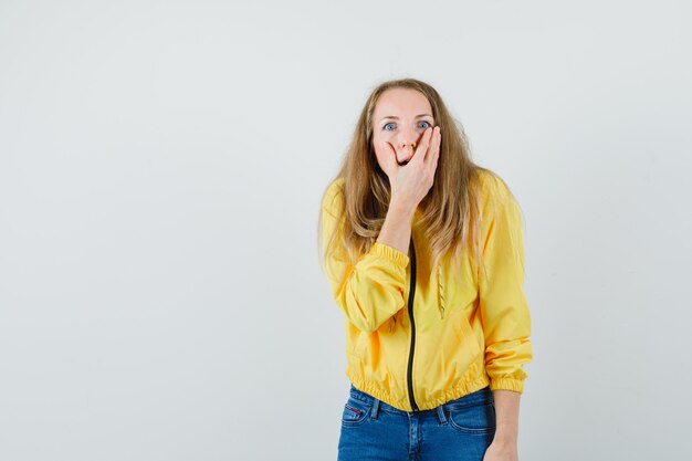 Young woman in yellow bomber jacket and blue jean covering her mouth and looking surprised , front view.