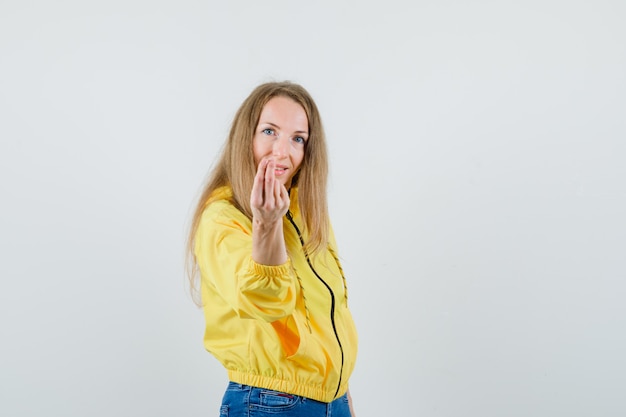 Young woman in yellow bomber jacket and blue jean inviting to come and looking optimistic , front view.