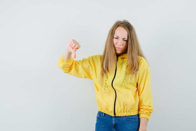 Young woman in yellow bomber jacket and blue jean showing thumb down and looking disleased , front view.