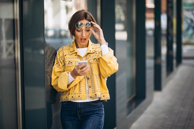 Free photo young woman in yellow jacket using phone outside in the street