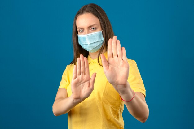 Young woman in yellow polo shirt and medical protective mask making stop gesture with hands on isolated blue background