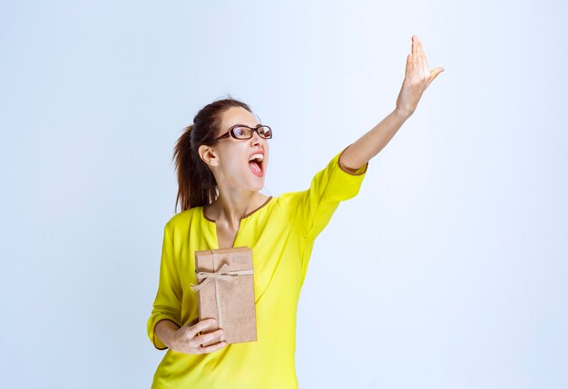 Young woman in yellow shirt holding a cardboard gift box and inviting a person to present it