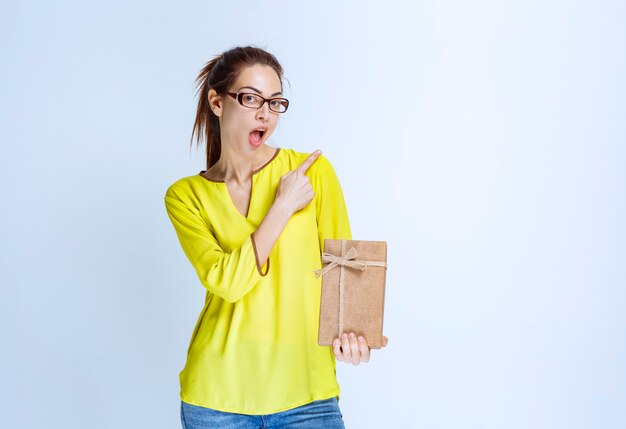 Young woman in yellow shirt holding a cardboard gift box and pointing at someone on the right