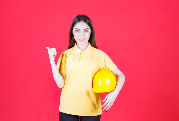 Free photo young woman in yellow shirt holding a yellow helmet and pointing at someone behind