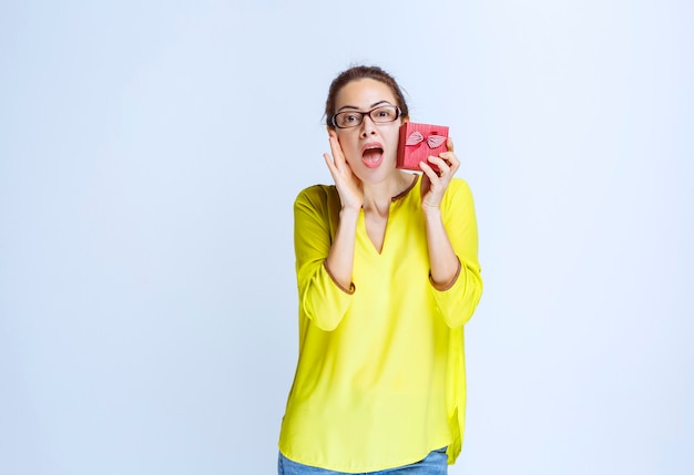 Young woman in yellow shirt showing her red gift box and looking surprized